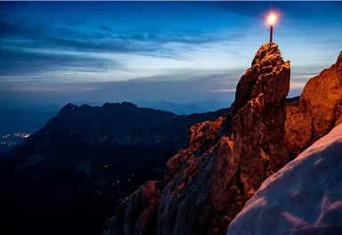 A picturesque mountain panorama at twilight with a burning candle on a rock. The scene shows snow-covered mountains and a clear sky in the background.