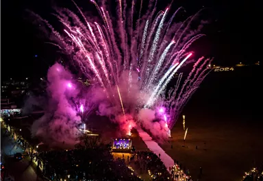 Ein spektakuläres Feuerwerk erleuchtet den Nachthimmel mit bunten Farben. Viele Menschen feiern und genießen die festliche Atmosphäre am Strand.