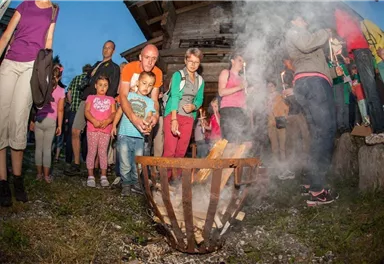 A group of people stands around a campfire and watches curiously. The smoke from the fire rises into the evening sky.