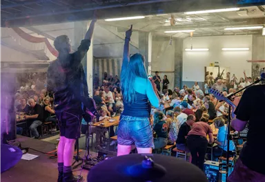 A band is playing on a stage in front of an enthusiastic audience. The spectators raise their hands and celebrate the music.