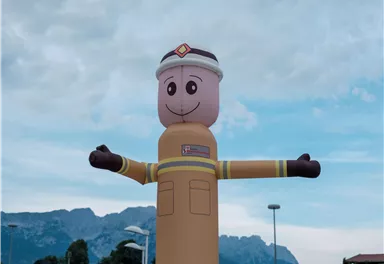 An inflatable firefighter with a friendly face stands with open arms. In the background, there are mountains and a cloudy sky.