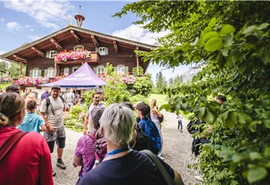 Eine Gruppe von Menschen steht vor einem traditionellen Holzhaus mit blühenden Fenstern. Im Hintergrund ist eine grüne Landschaft zu sehen, die den schönen Tag unterstreicht.