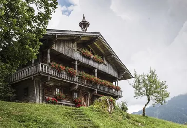 A traditional wooden house with a balcony and colorful flowers. It stands in a green landscape under a cloudy sky.
