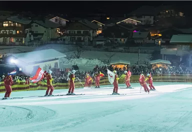 A skiing event at night with several skiers in red suits. In the background, illuminated houses and many spectators can be seen.