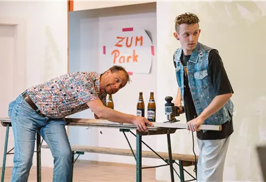 Zwei Männer stehen an einem Tisch in einem Raum. Einer beugt sich über den Tisch, während der andere eine Kamera hält und in die Ferne blickt.