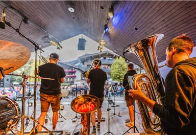 A band is playing on a stage in front of an audience. In the foreground, musicians with wind instruments can be seen.