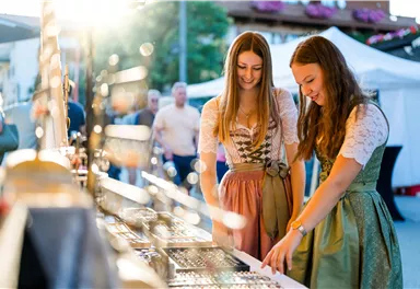 Two women in traditional dresses are looking at jewelry at a market. The sun is shining, and in the background, other visitors can be seen.