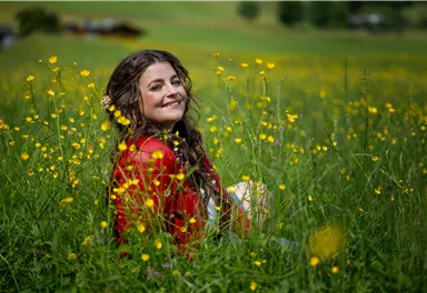 A smiling woman is sitting in a blooming field of yellow flowers. She is wearing a red dress and enjoying nature.