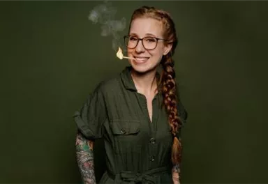 Eine lächelnde Frau mit langen, geflochtenen Haaren und Brille steht vor einem grünen Hintergrund. Sie hat Tattoos auf ihren Armen und raucht eine Zigarette.