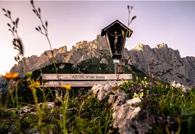 Eine malerische Berglandschaft mit einer Holztafel und einer kleinen Kapelle. Bunte Blumen wachsen im Vordergrund, während die imposing Felsen im Hintergrund sichtbar sind.