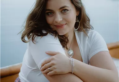 A person is sitting in a boat by the water and smiling at the camera. The surroundings are calm and natural, with soft colors.