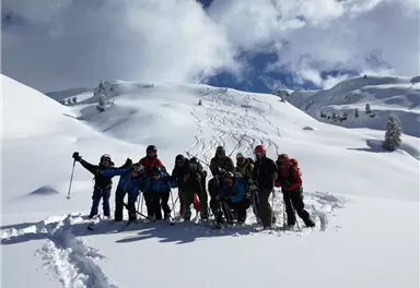 A group of skiers happily poses in the snow. In the background, snow-covered hills and a clear sky can be seen.
