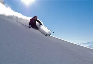 A skier glides down the snowy slope with momentum. The sky is clear and sunny, and fresh snow is being kicked up.