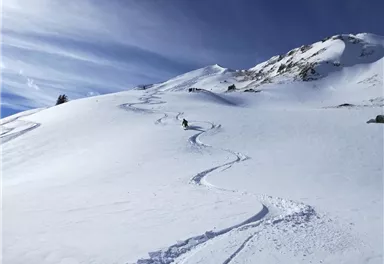 A snow-covered mountain landscape with a skier leaving a trail in the fresh snow. The sky is clear and blue, creating a beautiful winter scene.