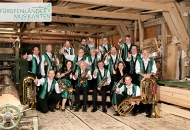 A group of musicians in traditional costume stands in a rustic setting. They hold instruments and smile at the camera.