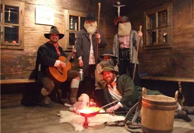 A group of five people in historical clothing stands in a rustic room. They pose around a fire in a wooden barrel and have various props.