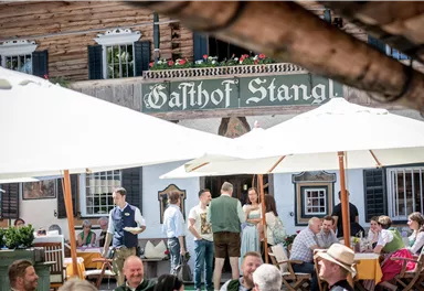 Eine belebte Terrasse eines Gasthauses mit Gästen unter Sonnenschirmen. Im Hintergrund ist das Schild "Gasthof Stangl" sichtbar.