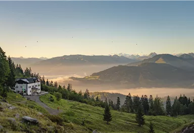 Eine malerische Berglandschaft mit grünen Wiesen und dichten Wäldern. Im Vordergrund steht ein gemütliches Haus, umgeben von sanften Hügeln und der Weite des Tals.