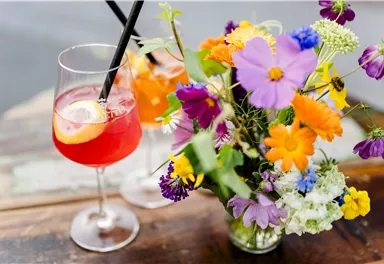 Ein bunter Blumenstrauß steht neben einem Cocktail mit einem roten Farbton und einer Zitronenscheibe. Die Atmosphäre ist fröhlich und einladend.