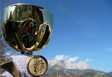 Ein goldener Pokal steht im Vordergrund, mit einer beeindruckenden Berglandschaft im Hintergrund. Der Himmel ist klar und blau.