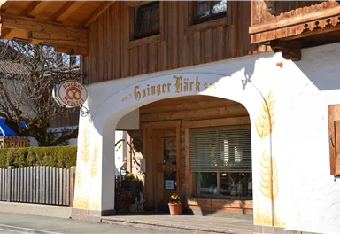 Ein charmantes Holzgebäude mit einem einladenden Eingang. Über der Tür steht "Ginger Bäck" und es gibt dekorative Elemente wie Ähren an der Wand.