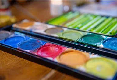 Eine Palette mit Wasserfarben in verschiedenen Farben. Im Hintergrund sind einige Malutensilien und eine unscharfe Umgebung zu erkennen.