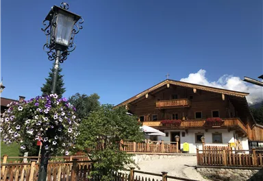 Ein traditionelles Holzhaus mit Balkon und schönen Blumen vor der Tür. Im Hintergrund ist ein blauer Himmel mit einigen Wolken zu sehen.
