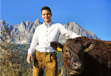 Ein junger Mann steht neben einem Rind in der Natur. Im Hintergrund sind beeindruckende Berge und ein blauer Himmel zu sehen.