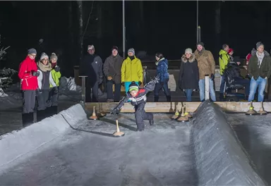 Eine Gruppe von Menschen beobachtet ein Kind, das auf einer Eisbahn spielt. Die Umgebung ist winterlich und es ist dunkel, mit nur wenigen Lichtern sichtbar.