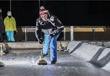 Ein Mann spielt Eisstockschießen auf einer beleuchteten Bahn. Im Hintergrund sind Zuschauer in Winterkleidung zu sehen.