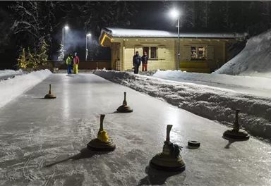 Eine Eisbahn, beleuchtet von Laternen, mit fünf Holzstöcken auf dem Eis. Im Hintergrund stehen mehrere Personen und eine kleine Hütte.