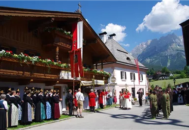 Eine traditionelle Feier in einem malerischen Dorf. Menschen in festlicher Tracht stehen zusammen, umgeben von historischen Gebäuden und Bergen.