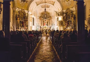 Eine stimmungsvolle Kirche mit vielen Menschen, die im Dunkeln sitzen. Der Raum ist festlich dekoriert und warm beleuchtet.