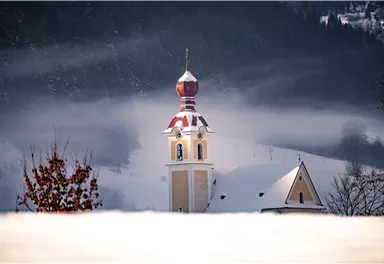 Ein Kirchturm mit einer roten Kuppel ragt aus einer schneebedeckten Landschaft hervor. Im Hintergrund sind verschneite Hügel und Nebel zu sehen.