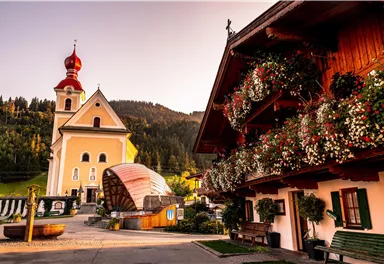 Ein malerisches Dorf mit einer Kirche und blühenden Balkonen. Im Hintergrund sind sanfte Hügel und Bäume zu sehen.