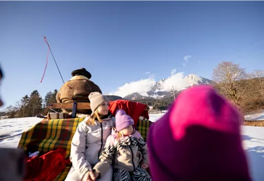 Eine Familie sitzt auf einem Schlitten im Schnee, umgeben von einer winterlichen Landschaft. Im Hintergrund sind Berge unter einem klaren blauen Himmel zu sehen.