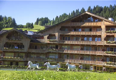 Ein charmantes Holzgebäude mit mehreren Balkonen in einer grünen Umgebung. Vor dem Gebäude laufen einige weiße Pferde über die Wiese.