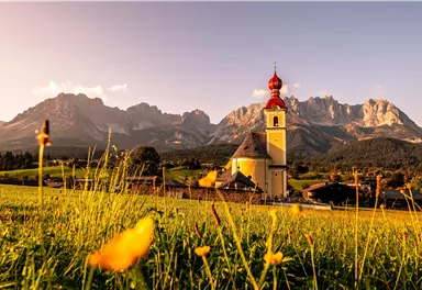 Eine malerische Kirche steht in einer grünen Wiese vor majestätischen Bergen. Die Landschaft ist von bunten Blumen und warmem Sonnenlicht geprägt.
