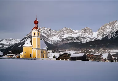 Eine bunte Kirche mit rotem Kirchturm steht in einer schneebedeckten Landschaft. Im Hintergrund sind beeindruckende Berge sichtbar.