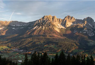 Eine majestätische Berglandschaft mit schroffen Gipfeln und grünen Wäldern im Vordergrund. Der Himmel ist klar und verleiht der Szene ein warmes Licht.