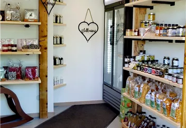 Ein Laden mit verschiedenen Regalen voller Lebensmittel und Getränken. Auf der linken Seite sind Gläser und Flaschen ausgestellt, während rechts ein Kühlschrank und weitere Waren stehen.