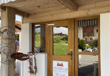 Der Eingang zu einem charmanten Geschäft mit Holzverkleidung und einem weißen Sonnenschirm. Auf der Tür hängt ein Schild mit dem Namen "Kaiser Ladl".