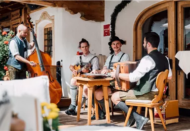 Eine Gruppe von Musikern spielt traditionelle Instrumente in einem gemütlichen Außenbereich. Die Musiker tragen bayerische Trachten und umgeben sich mit schöner Dekoration.