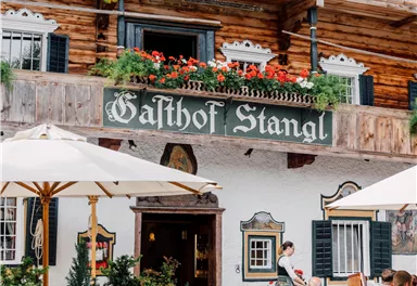 Ein traditionelles Gasthaus mit holzverkleideter Fassade und blühenden Pflanzen. Tische im Freien unter Sonnenschirmen bieten einen einladenden Platz zum Verweilen.