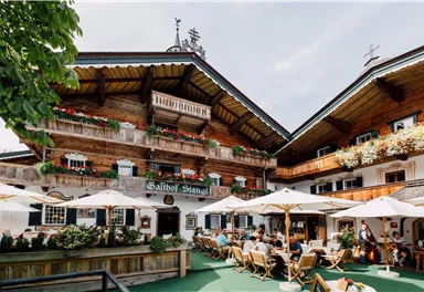 Ein traditionelles alpenländisches Gasthaus mit Holzfassade und blühenden Balkonen. Gäste sitzen unter Sonnenschirmen im Freien und genießen die Atmosphäre.