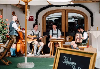 Eine Gruppe von Musikern in traditioneller Tracht spielt in einem gemütlichen Außenbereich. Die Atmosphäre ist entspannt und einladend.