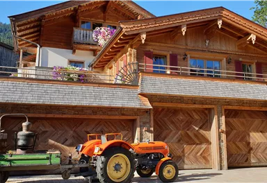 Ein traditionelles Holzhaus mit einem Balkon und Blumen. Vor dem Haus steht ein oranger Traktor und ein Wagen mit Blumen.