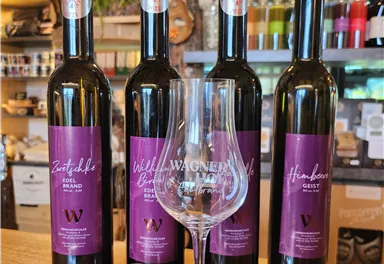 Vier Flaschen mit Wein, die nebeneinander stehen, und ein Weinglas im Vordergrund. Die Etiketten sind in lila gehalten und enthalten verschiedene Namen.