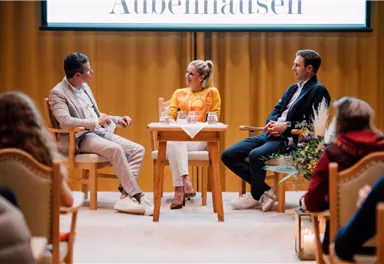Eine Panel-Diskussion mit drei Personen auf einer Bühne. Im Hintergrund ist ein großes Banner mit dem Wort "Außenmalen" zu sehen.