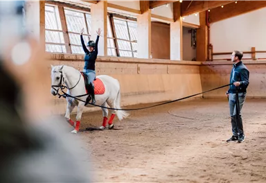 Eine Reiterin zeigt eine Akrobatikübung auf einem weißen Pferd in einer Reitarena. Der Trainer steht daneben und beobachtet die Übung.
